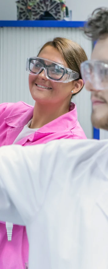 Female student in pink lab coat holding up flask while male student focuses on pipette Female student in pink lab coat holding up flask while male student focuses on pipette